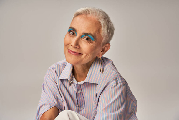 senhora madura positiva com maquiagem ousada em camisa listrada azul sorrindo para a câmera em cinza, banner - Foto, Imagem