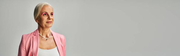 verträumte und stylische reife Dame in rosa Blazer und Perlenkette mit Blick auf Grau, Banner - Foto, Bild