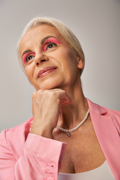 retrato de modelo senior de ensueño en blazer rosa sosteniendo la mano cerca de la barbilla y mirando hacia otro lado en gris - Foto, Imagen