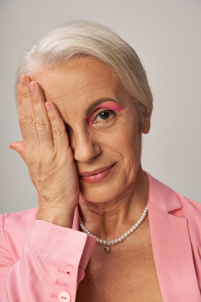 portrait of elegant mature lady in pearl necklace and pink blazer obscuring face with hand on grey - Photo, Image