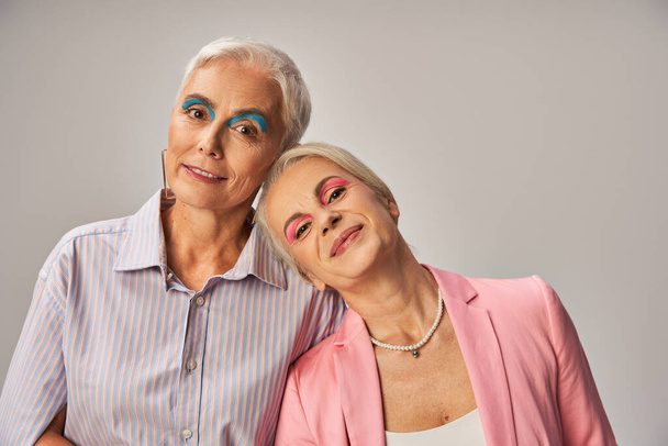 радісні старші друзі-жінки в модному синьо-рожевому вбранні дивиться на камеру на сірому, щасливому - Фото, зображення