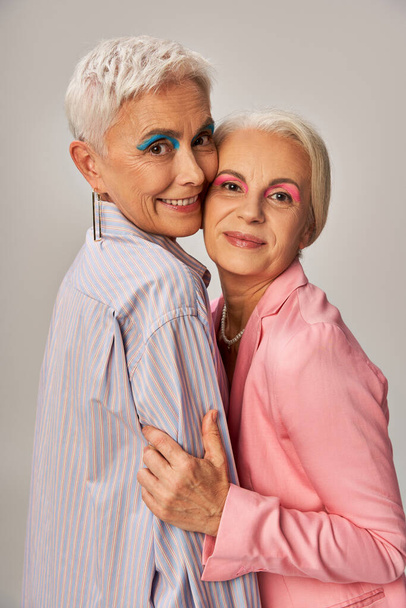 χαρούμενη ηλικιωμένη fashionistas σε μπλε και ροζ ρούχα αγκαλιάζοντας και κοιτάζοντας την κάμερα σε γκρι φόντο - Φωτογραφία, εικόνα