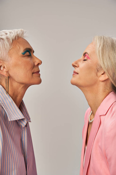 elegantes amigas mayores en ropa azul y rosa de pie con los ojos cerrados en gris, vista lateral - Foto, imagen
