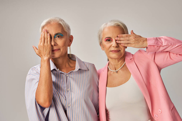 módní starší modely v modrém a růžovém opotřebení zakrývaly tváře rukama a dívaly se na kameru na šedé - Fotografie, Obrázek