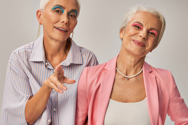 модная пожилая женщина с синей подводкой для глаз, указывающая пальцами на камеру рядом с веселой подругой на серой - Фото, изображение