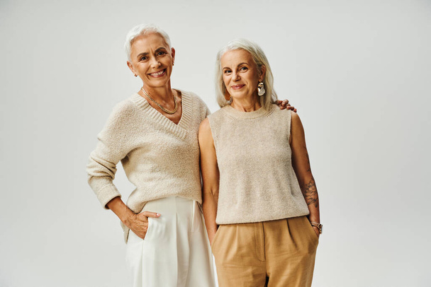 élégant vieillissement, joyeuses amies en tenue décontractée tendance posant avec les mains dans les poches sur gris - Photo, image
