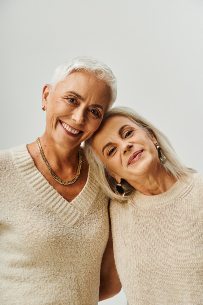 Porträt lächelnder Seniorinnen in Make-up und goldenen Accessoires, die vor grauem Hintergrund in die Kamera schauen - Foto, Bild