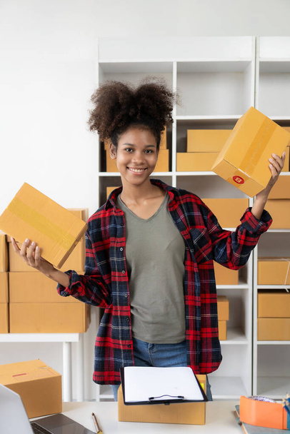Small business startup SME app African female business owner using laptop receives and verifies online orders to prepare product boxes and ship them. vertical image - Foto, Imagen