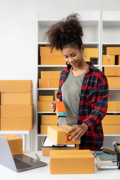 Small business startup SME app African female business owner using laptop receives and verifies online orders to prepare product boxes and ship them. vertical image - Фото, зображення