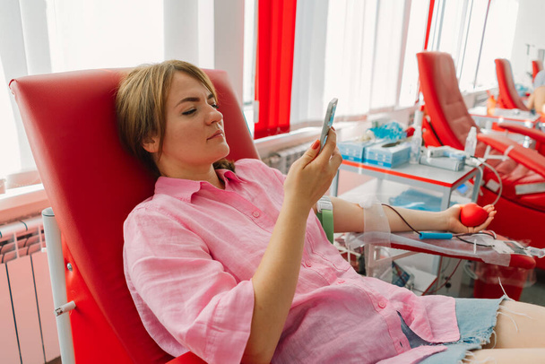 Молода жінка з телефоном робить донорство крові в лікарні. Жінка пожертвувала кров, тримаючи червоне серце. Донорство крові. - Фото, зображення