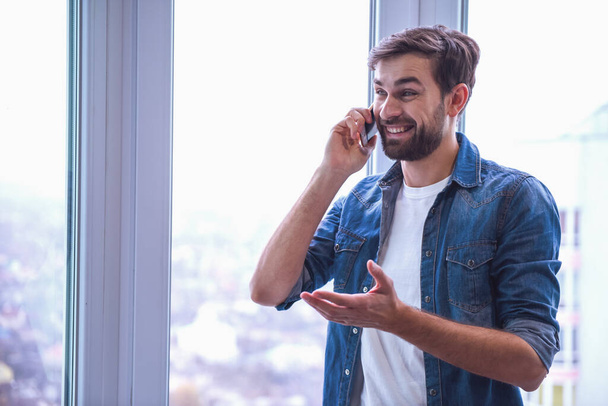 Красивый мужчина в джинсовой одежде разговаривает по мобильному телефону и улыбается, стоя у окна
 - Фото, изображение