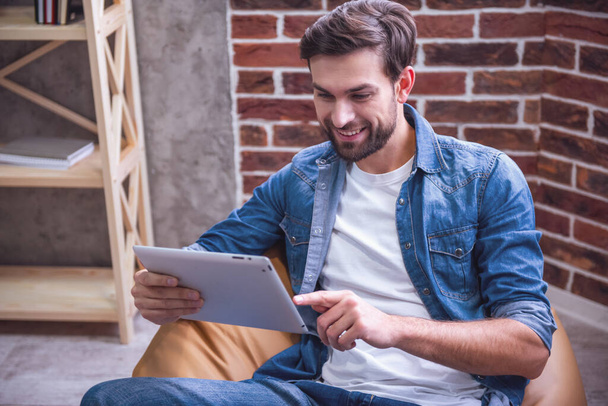Pohledný muž v jean oblečení používá digitální tabletu a usmívající se přitom sedí na gauči - Fotografie, Obrázek