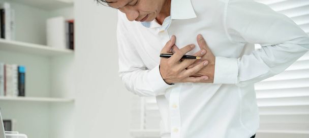 torace mano con sintomi di attacco di cuore, l'uomo asiatico che lavora duramente ha dolore al petto causato da malattie cardiache, perdita, dilatazione, ingrossamento del cuore coronarico, premere sul petto con un'espressione dolorosa - Foto, immagini