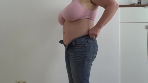 Неузнаваемая пухленькая женщина в джинсах небольшого размера, розовый топ, плюс грудь размера, с толстым животом, обвисшим, сжимая лишний жир на талии. Попытка надеть маленькие джинсы. - Кадры, видео
