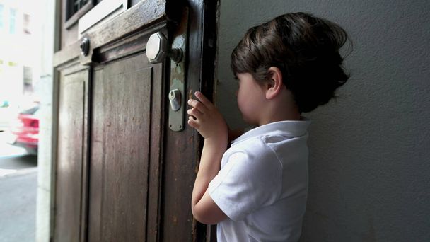 Kleine jongen die de voordeur opent en buiten stapt in de straat. Kind reikt naar deurknop en verlaat appartement, stappen op de stoep - Foto, afbeelding