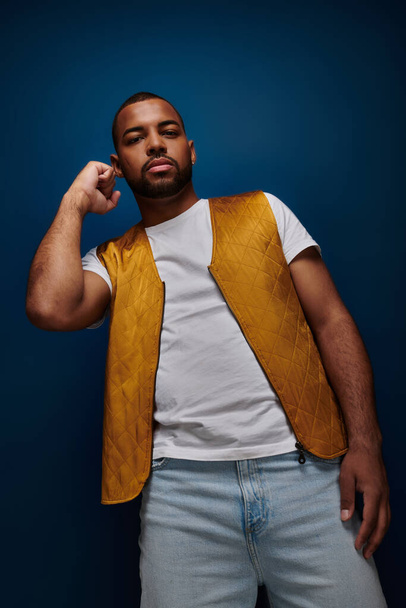 handsome stylish man in yellow vest posing with raised fist on dark blue backdrop, fashion concept - Photo, Image
