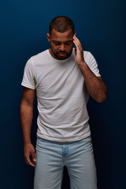 молодой человек в стильной одежде позирует на темно-синем фоне с рукой возле лба, концепция моды - Фото, изображение