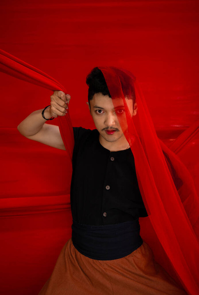 азіатський чоловік у чорній сорочці, що закриває обличчя червоною тканиною з переляканим виразом обличчя перед червоним тлом - Фото, зображення