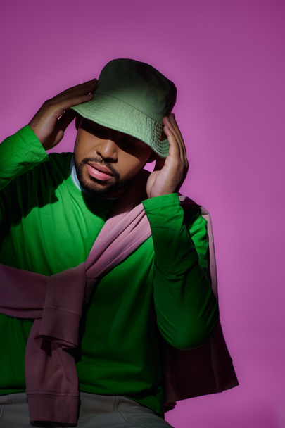 stylowy Afroamerykanin człowiek w zielonej bluzie dotykając jego panama na fioletowym tle, moda - Zdjęcie, obraz