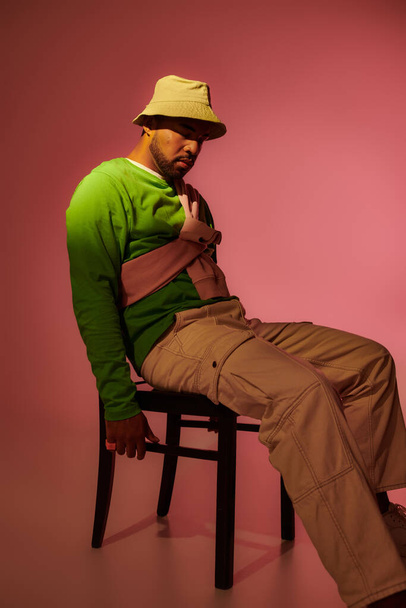 good looking african american man in green sweatshirt and panama sitting on chair, fashion concept - Photo, Image
