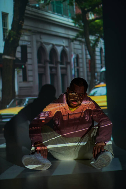 pohledný stylový muž v neformálním oblečení sedící na podlaze v digitálních projektorových světlech, módní koncept - Fotografie, Obrázek