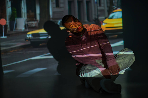 joven afroamericano hombre reclinado ligeramente en el suelo con las manos en las piernas cruzadas, concepto de moda - Foto, Imagen