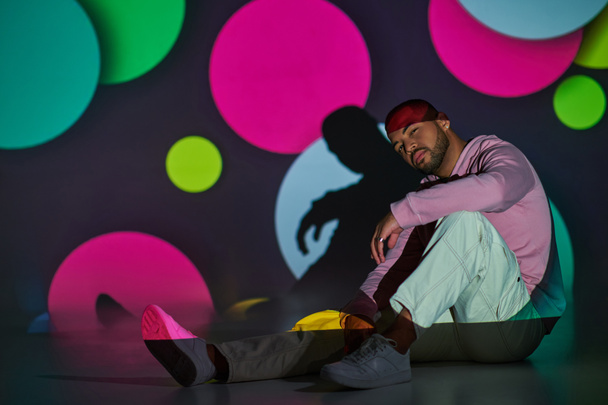 pohledný mladý muž sedící na podlaze, lehce se usmívající v digitálních projektorových světlech, módní koncept - Fotografie, Obrázek