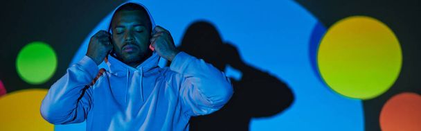 νεαρός άνδρας που βάζει κουκούλα με κλειστά μάτια σε ψηφιακά φώτα προβολέα, έννοια της μόδας, πανό - Φωτογραφία, εικόνα