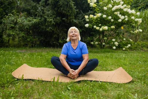 Mujer mayor atlética de los años 50 haciendo poses en la esterilla de yoga al aire libre vestida con ropa deportiva sola en el parque o bosque en la naturaleza. Foto de alta calidad - Foto, imagen