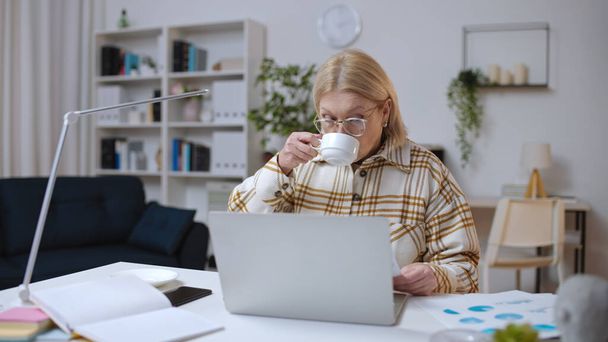 Mature woman working with business charts and drinking tea in a home office - Foto, Bild