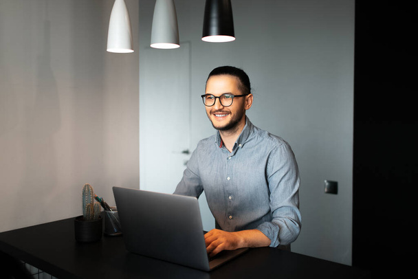 Portret van een knappe, jonge gelukkige man, die thuis werkt op een laptop. Draag een bril en een T-shirt. Achtergrond van de grijze muur. - Foto, afbeelding