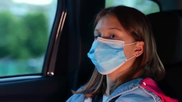 Vidám kislány védő maszkban, aki elfordítja a tekintetét, miközben a hátsó ülésen ül az autóban az utazás alatt. Utazás, szállítás, gyerekkori koncepció. Valós idejű - Felvétel, videó