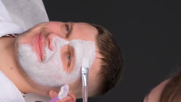 Молодой человек в спа, проходящий процедуру по уходу за лицом. Глиняная маска на лице человека. Косметолог выполняет массаж лица с увлажняющей маской, нанесенной на лицо. Вертикальное видео - Кадры, видео