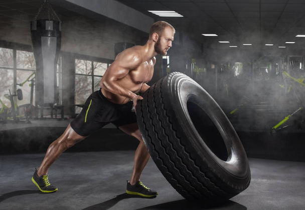 Young adult man flipping and rolling tire during exercise in the gym. Sports concept, fat burning and a healthy lifestyle. - Photo, Image