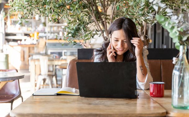 Bir kafede dizüstü bilgisayarla çalışan bir kadın telefonda konuşuyor. Teknoloji, iş ve serbest meslek kavramı. - Fotoğraf, Görsel