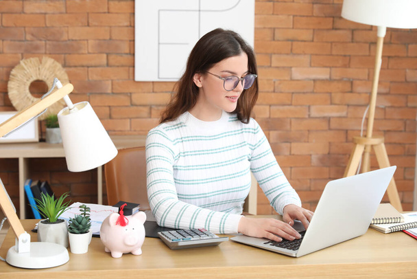 Joven estudiante sentada en la mesa con laptop y calculando sus finanzas. Concepto de préstamo estudiantil - Foto, Imagen