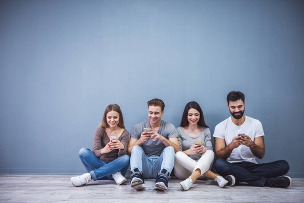 Jovens bonitos estão usando smartphones e sorrindo enquanto estão sentados no chão
 - Foto, Imagem