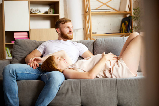 Coziness and care. Husband spending time with his beautiful pregnant wife at home. Woman lying on mans leg. Concept of pregnancy, family, love, relationship, parenthood and childhood - Valokuva, kuva