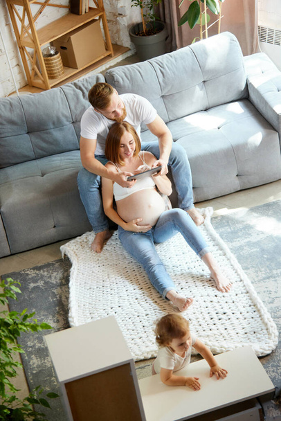 Happy, loving couple, man and pregnant woman sitting on couch at home and spending time together with little daughter. Concept of pregnancy, family, love, relationship, parenthood and childhood - Foto, afbeelding