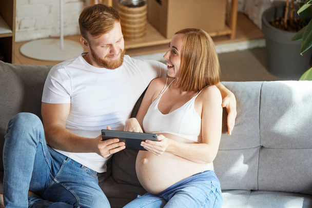 Young couple, future parents sitting at home and looking on tablet. Expecting baby, learning information. Concept of pregnancy, family, love, relationship, parenthood and childhood - Photo, image