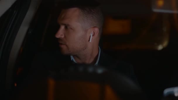 Fehér férfi vezeték nélküli fülhallgatóban, aki este a városban vezet autót. Jobb kormányos kocsi. Utazás, utazás, éjszakai élet koncepció. Valós idejű - Felvétel, videó