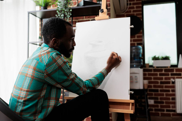 Jeune Afro-Américain s'exprimant à travers l'art, suivant des cours de dessin, étudiant masculin améliorant les compétences de croquis. Artiste talentueux assis au chevalet créer chef-d'œuvre sur toile - Photo, image