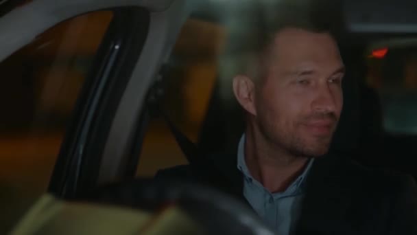 Un gars souriant conduisant et regardant le rétroviseur en voiture la nuit. Feux de circulation derrière l'homme. Voiture conduite à droite. Voyager, voyager, concept de vie nocturne. Temps réel - Séquence, vidéo