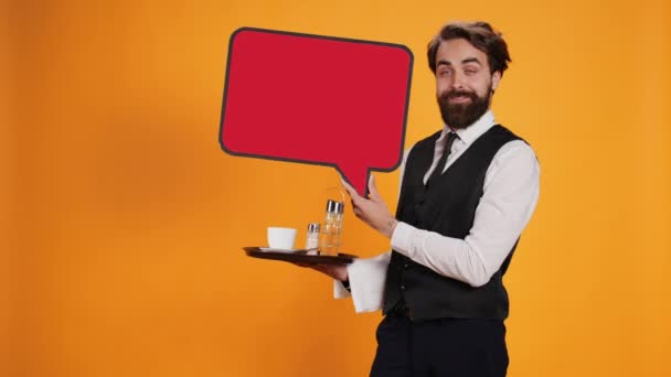 Elegant waiter holds speech bubble icon on camera, working in restaurant industry to create marketing ad with red cardboard cutout. Employee presenting billboard with blank isolated space. - Footage, Video