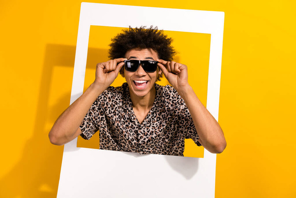 Photo of optimistic impressed guy with afro hair dressed print shirt touching sunglass in frame isolated on yellow color background. - Foto, afbeelding