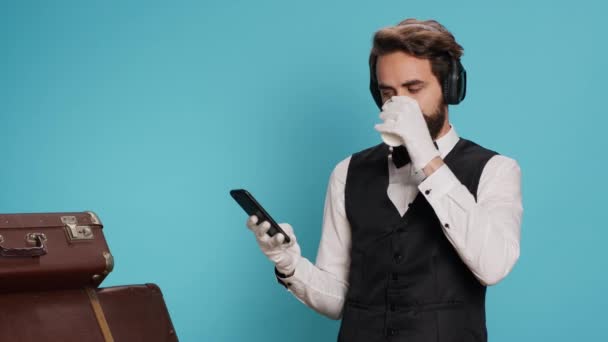 Doorman texting messages drinking coffee in studio, enjoying break with music on headphones. Young adult with classy hotel sector job messaging and sipping on cold brew beverage. - Footage, Video