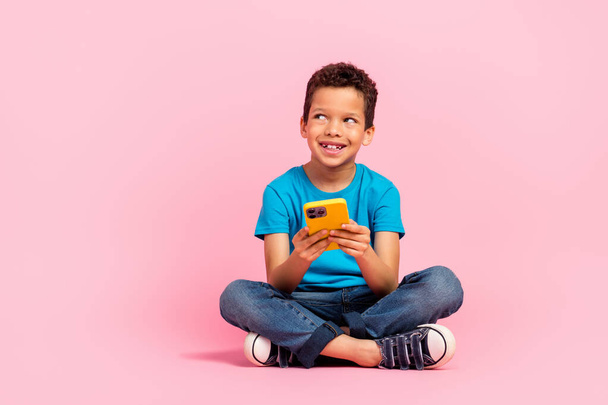 Full length photo of excited dreamy small boy wear blue t-shirt texting modern gadget looking empty space isolated pink color background. - 写真・画像