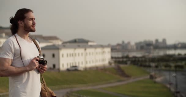 Zdjęcie w czasie rzeczywistym młodego brodatego mężczyzny o długich włosach, stojącego na widoku i obserwującego miasto, a następnie robiącego zdjęcia na kamerze filmowej retro przed zamazanym krajobrazem miasta podczas podróży - Materiał filmowy, wideo