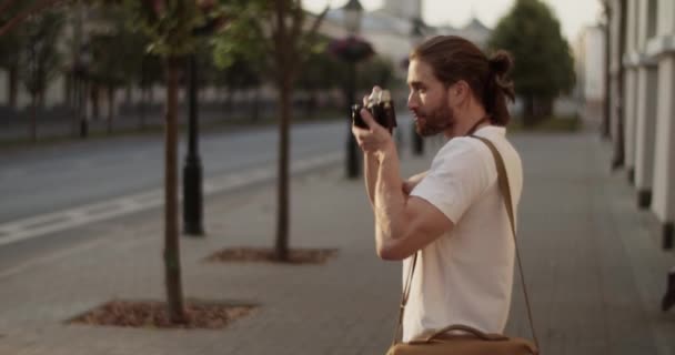 Boční pohled zoom mladého vousatého dlouhosrstého turisty s hnědou taškou na rameni stojící na ulici města a fotografování starého města na retro filmové kameře - Záběry, video