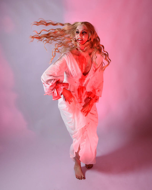 Full length portrait scary vampire zombie bride wearing bloody halloween fantasy costume dress. Dynamic jumping pose gestural arms reaching out. Isolated  studio backdrop with cinematic red lighting. - Фото, зображення